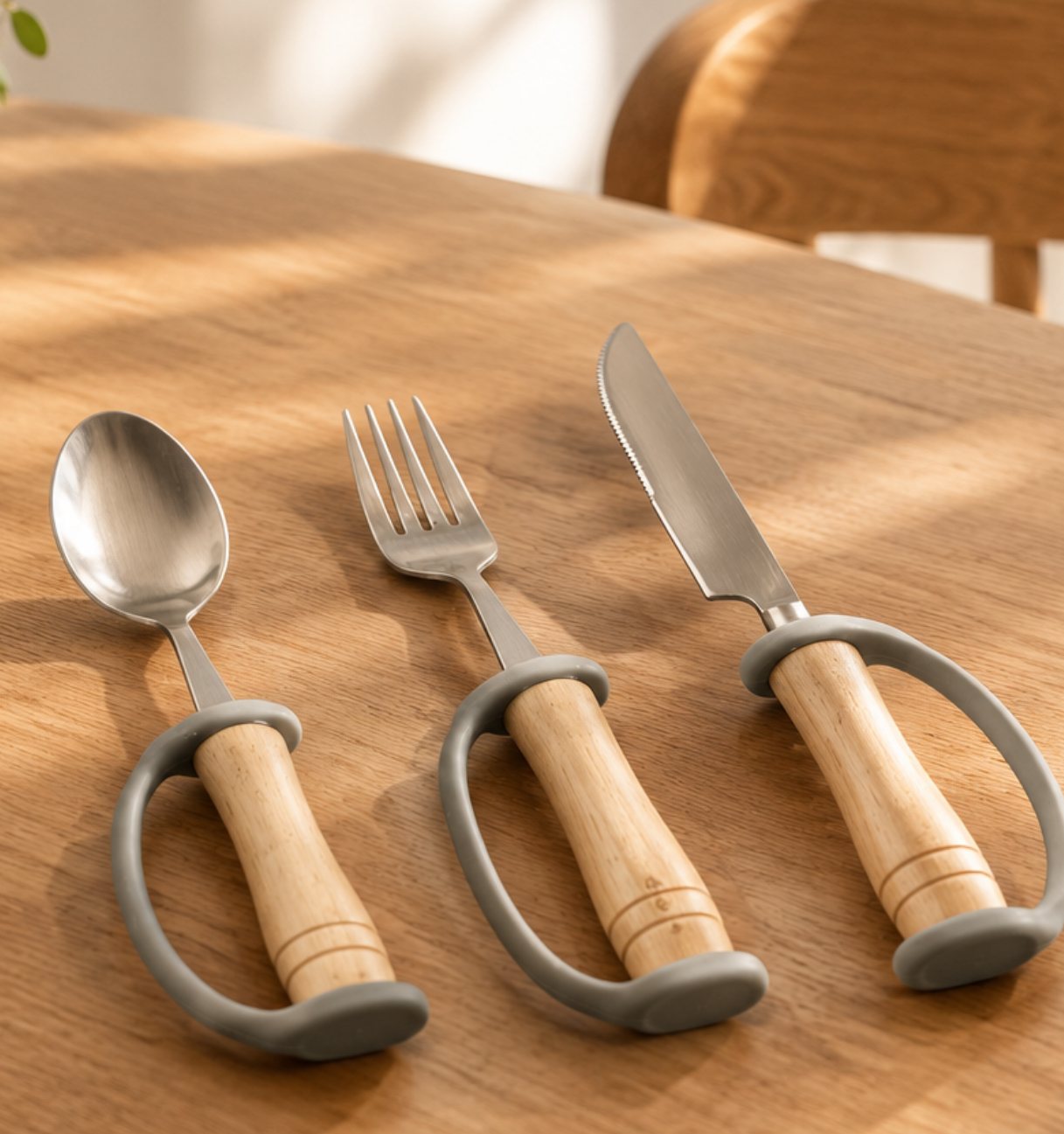 A spoon, fork and knife with turned timber handles and soft grey looped supports, laid side by side on a timber dining table.