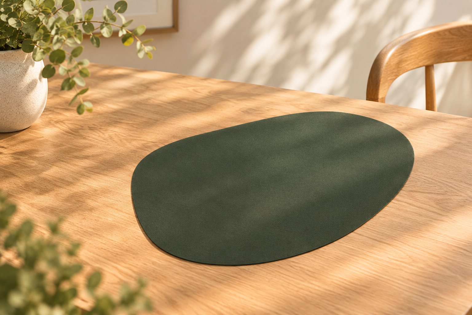 A forest green non-slip placemat with a soft, rounded pebble shape on a sunlit timber table.