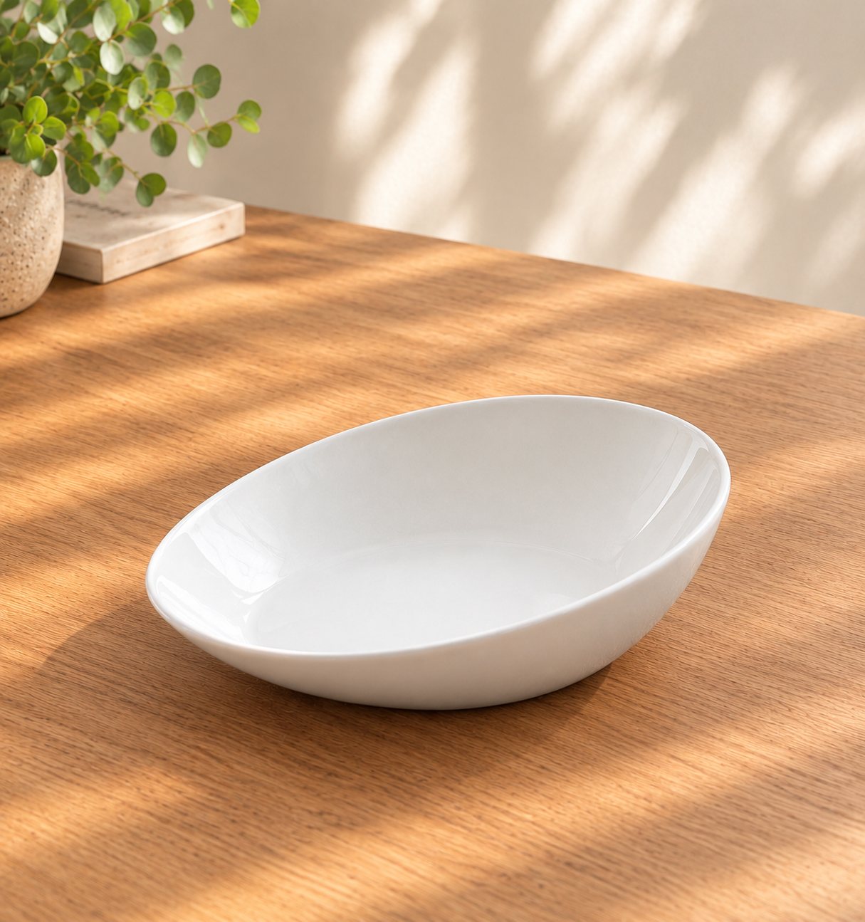 A white porcelain scoop plate with a sloped base and raised, curved edge on a sunlit timber table.
